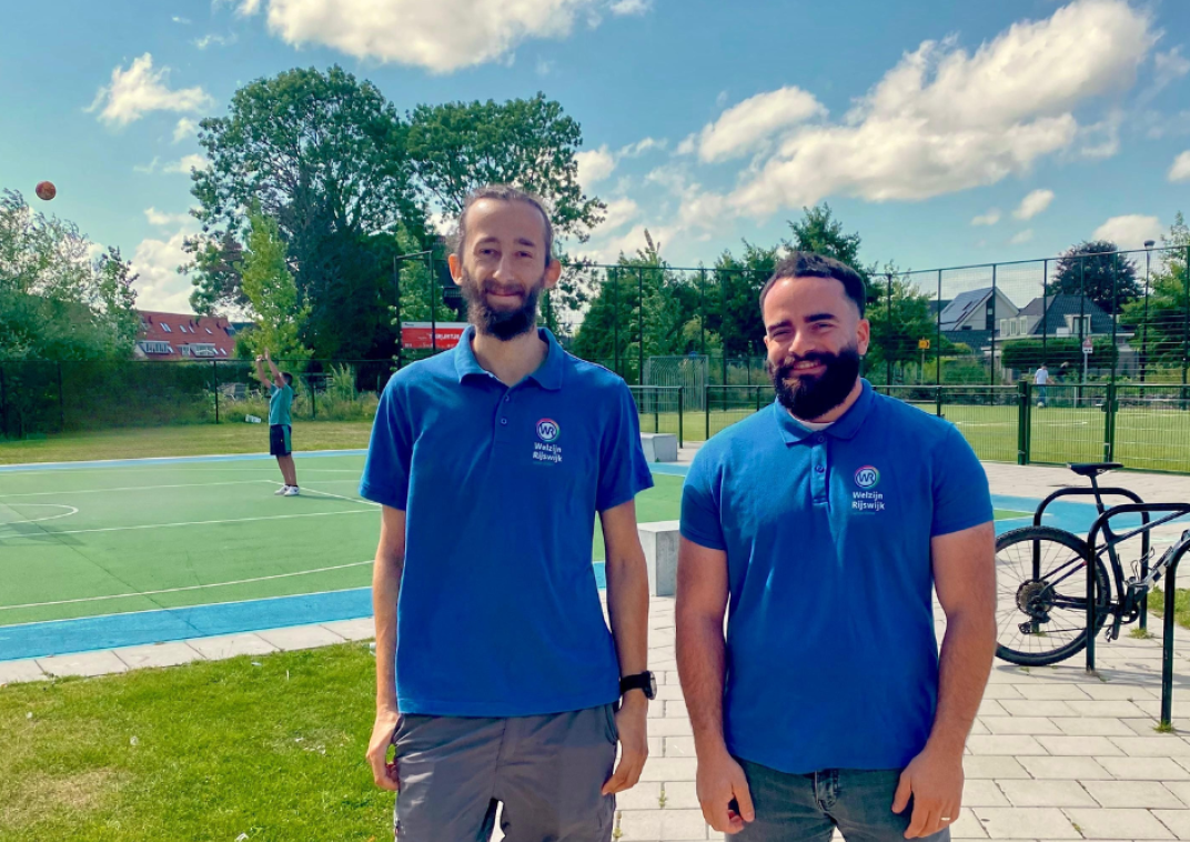 Twee mannen in blauwe werkkleding staan voor een sportachtige omgeving