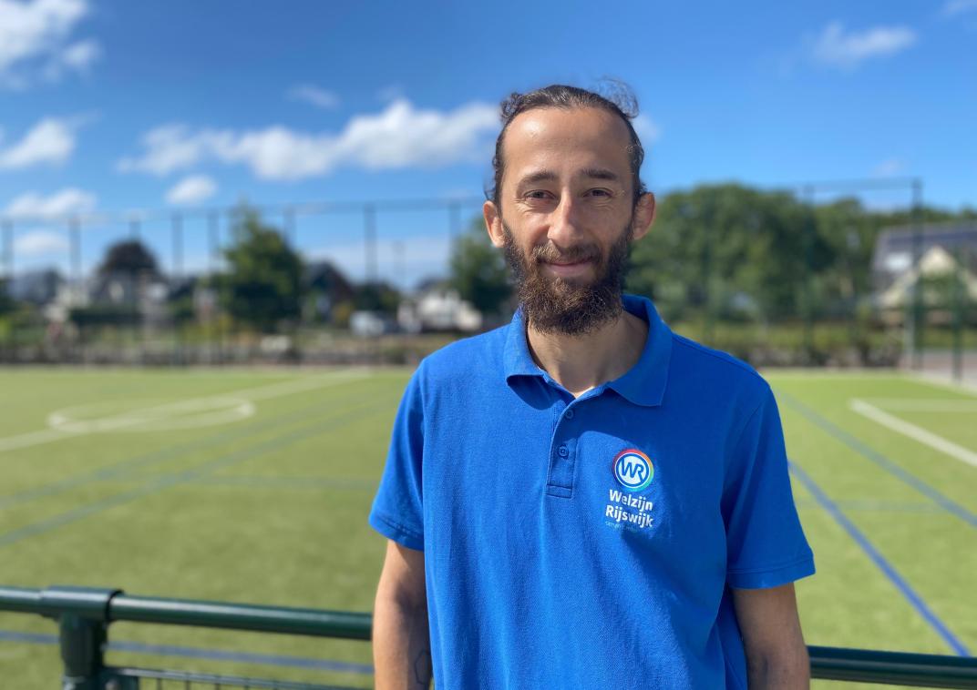 Portret van een man in een blauw shirt die voor een sportveld staat.