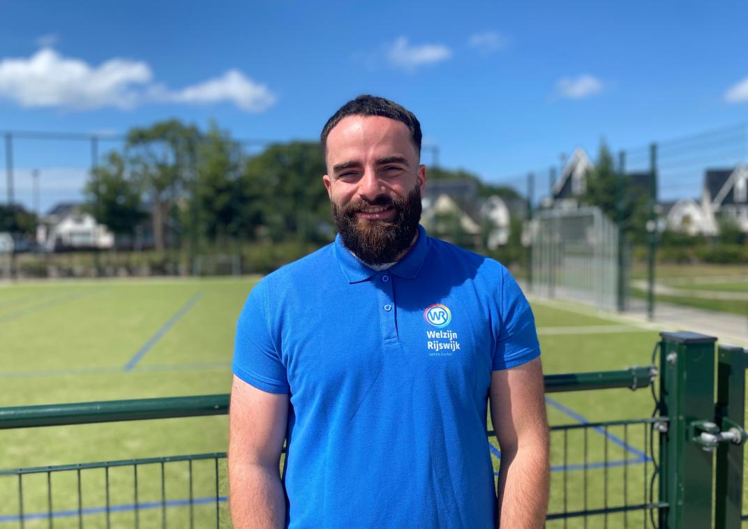 Man in blauwe kleding voor een hek met daarachter een voetbalveld