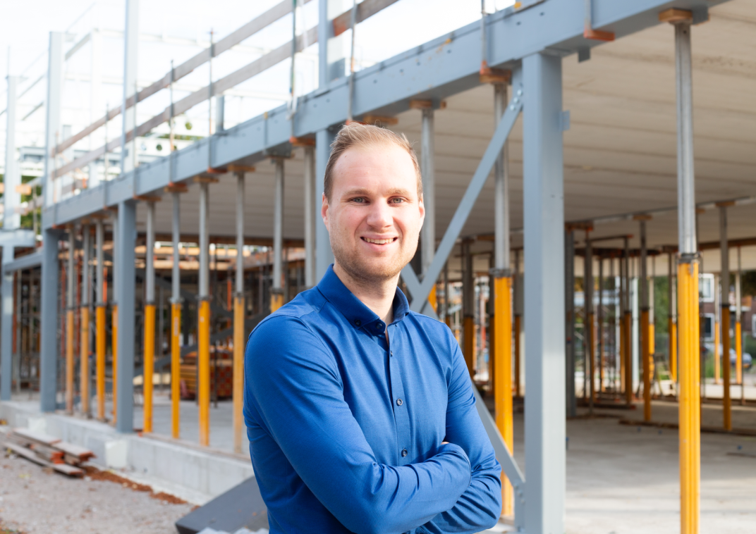 Man staat met zijn armen over elkaar en lacht naar de camera. Achter hem zie je het geraamte van een school die in aanbouw is. 