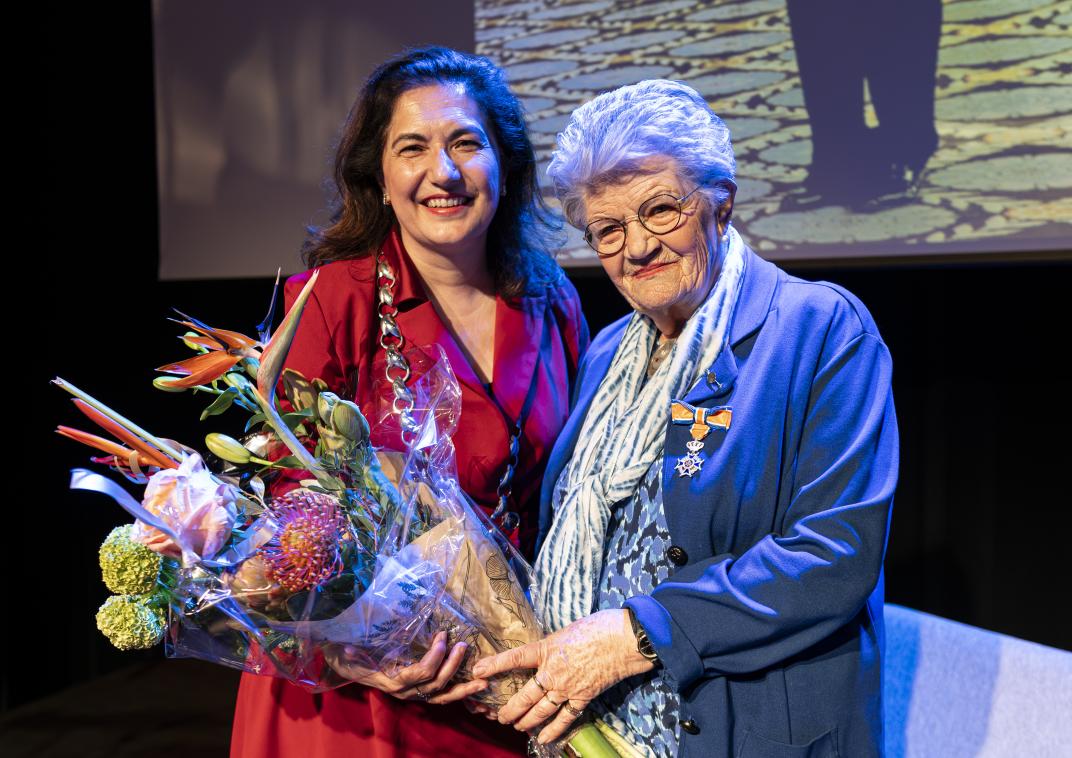 twee vrouwen kijken blij, een van de vrouwen draagt een bos bloemen en heeft een lintje opgespeld