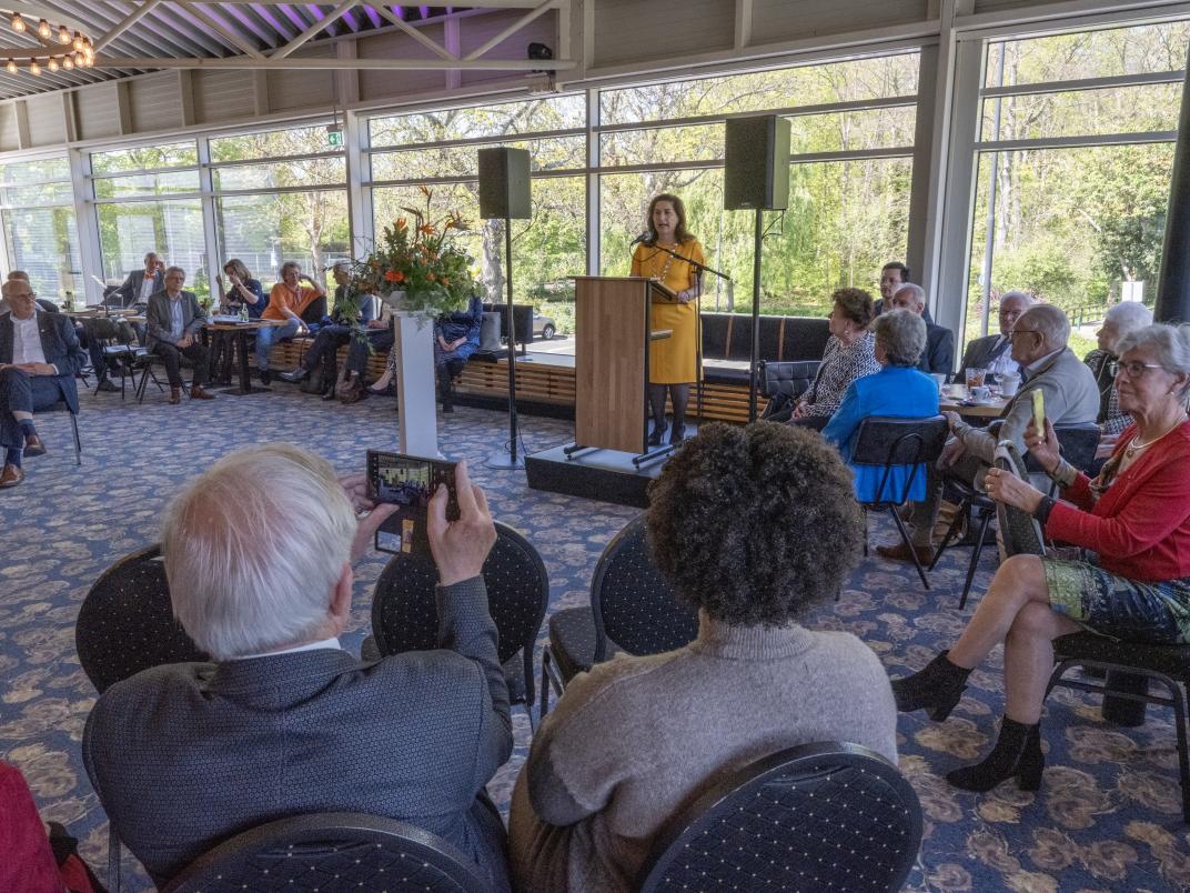 Burgemeester geeft een speech in een ruimte gevuld met mensen die aandachtig naar haar luisteren. Zij zitten aan tafels rondom het kleine podium.