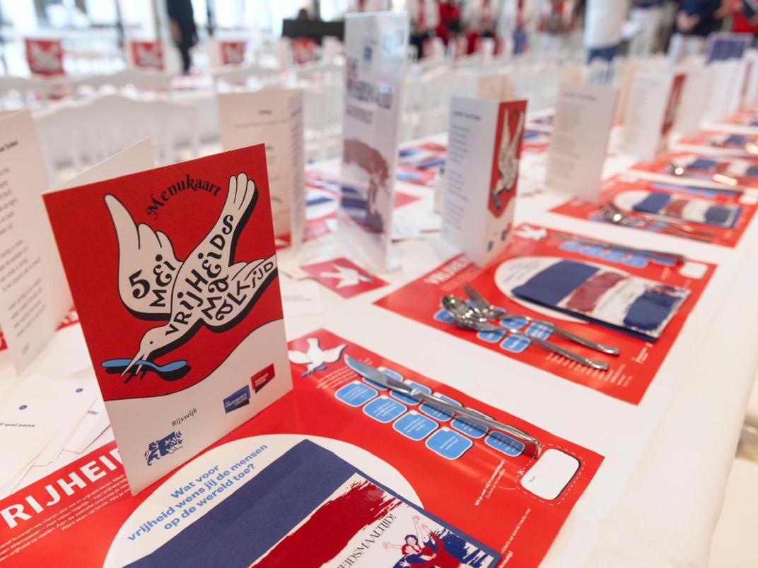 Lange tafel gedekt met placemet en een kaart in stijl van de vrijheidsmaaltijd. De kleuren zijn rood, wit en blauw en op de kaart staat een grote witte duif.