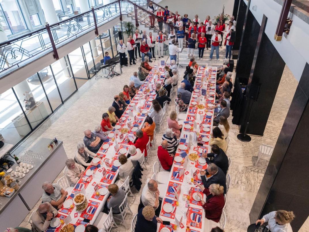 Foto van bovenaf genomen van 2 lange gedekte tafels met zo'n 60 mensen. Zij kijken naar een optreden van een koor terwijl zij genieten van een lunch.
