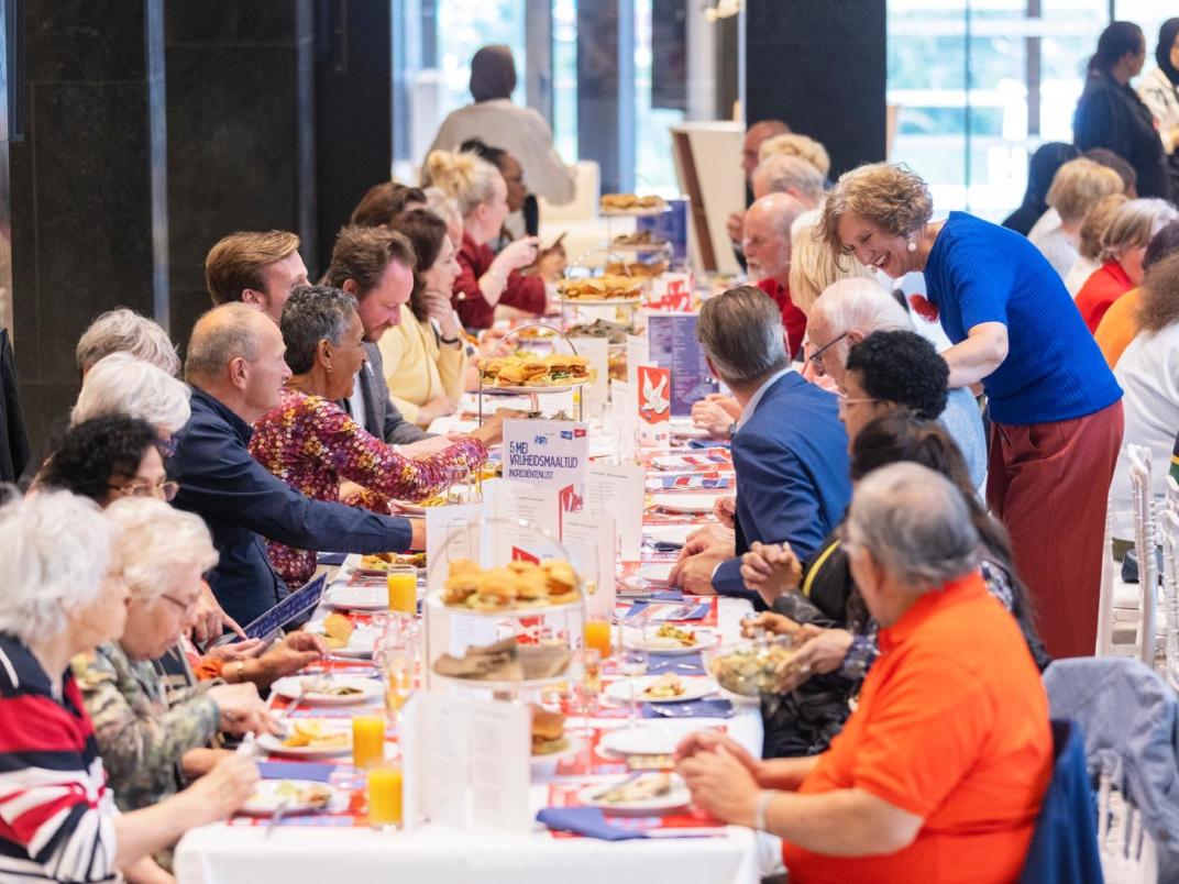 Een tafel gevuld met lunch producten. Oudere mensen aan tafel genieten van de lunch en praten met elkaar.