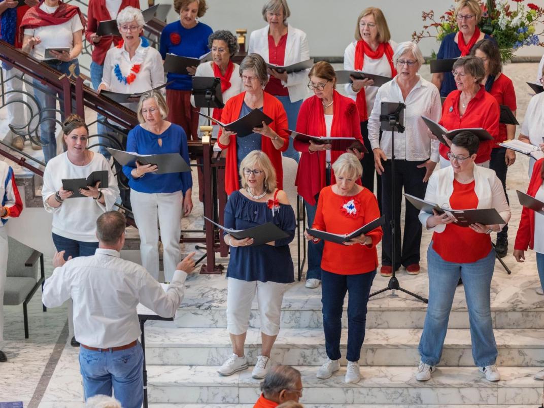 Zo'n 15 personen van een koor staan in groepsvorm met elkaar te zingen. Ze dragen kleding in rood, wit of blauwe kleuren.