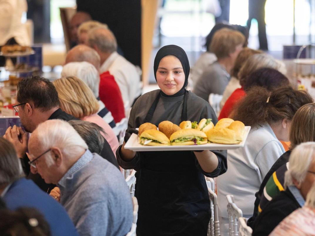 Leerling serveert een plateau met belegde broodjes uit. Om haar heen zitten oudere mensen aan twee lange tafels met elkaar te lunchen. De leerling staat centraal op de foto. 