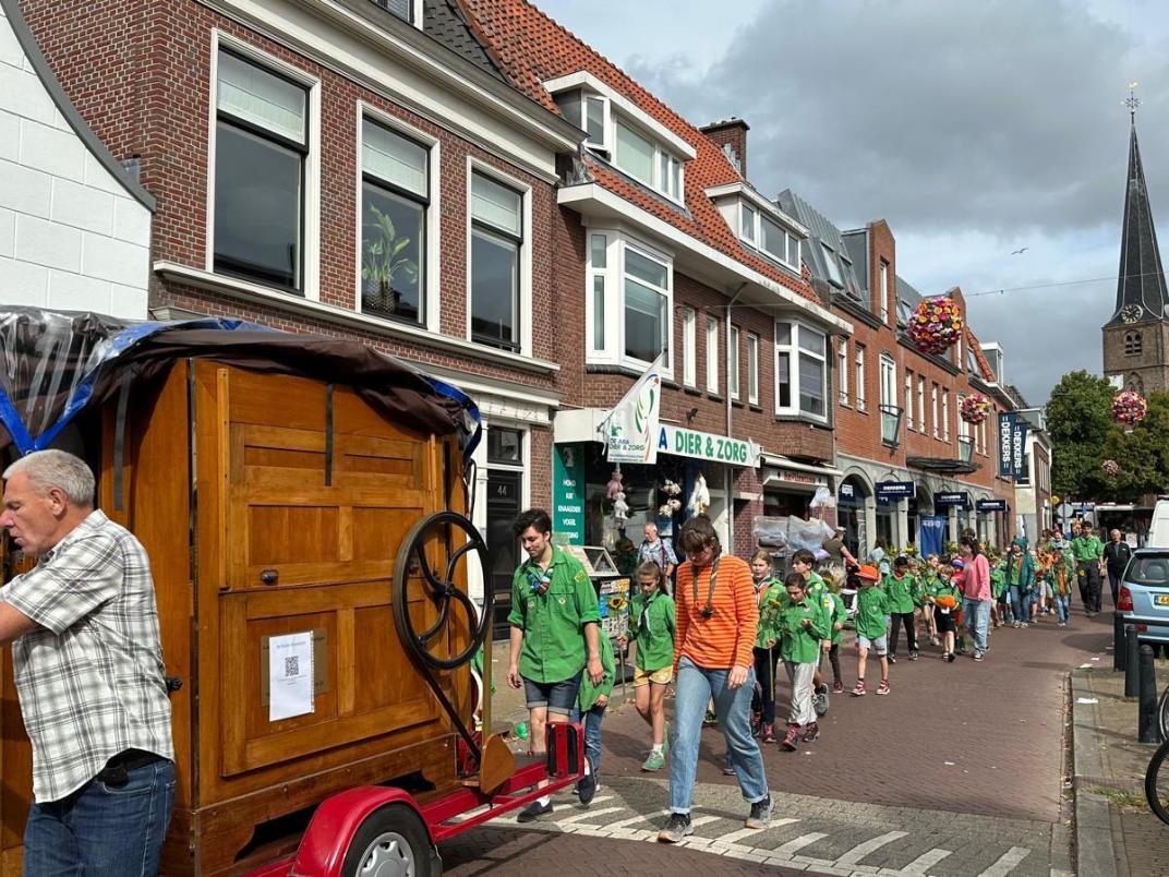 Kinderen lopen door een straat met huizen achter een man met een draaiorgel
