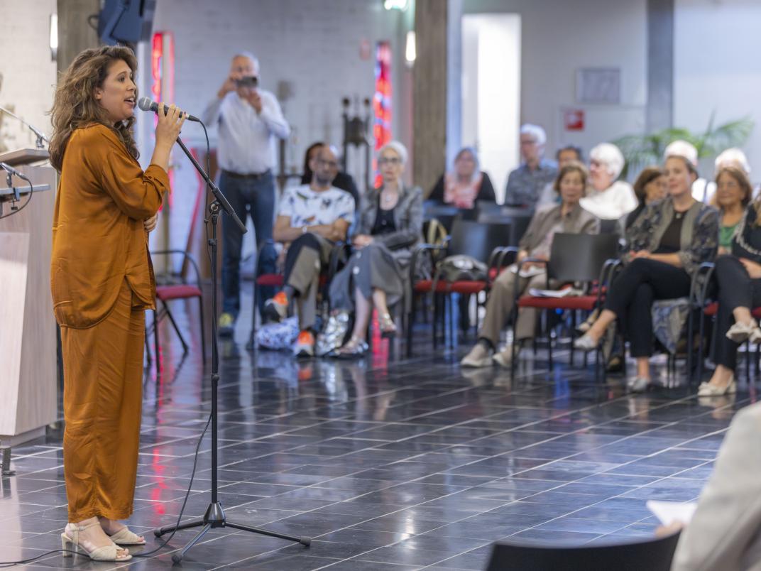 Een vrouw staat met een microfoon in haar hand en spreekt het publiek toe