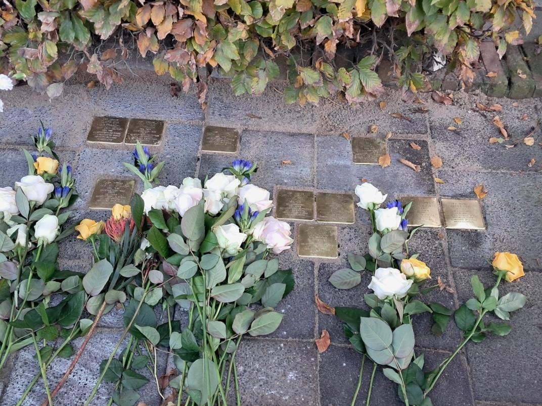 Witte, gele en paarse bloemen liggen rondom de gouden struikelstenen.