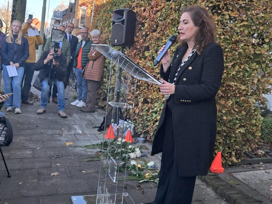 De burgemeester geeft een speech tijdens de herdenking. Ze heeft een microfoon in haar hand en draagt zwarte kleding.