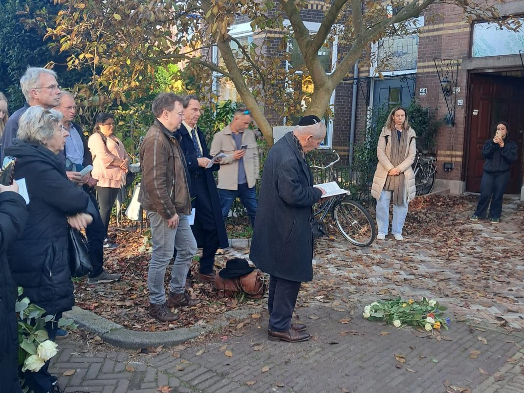 Verschillende mensen staan bij de struikelsteen. Een man leest hierbij voor uit een boek.