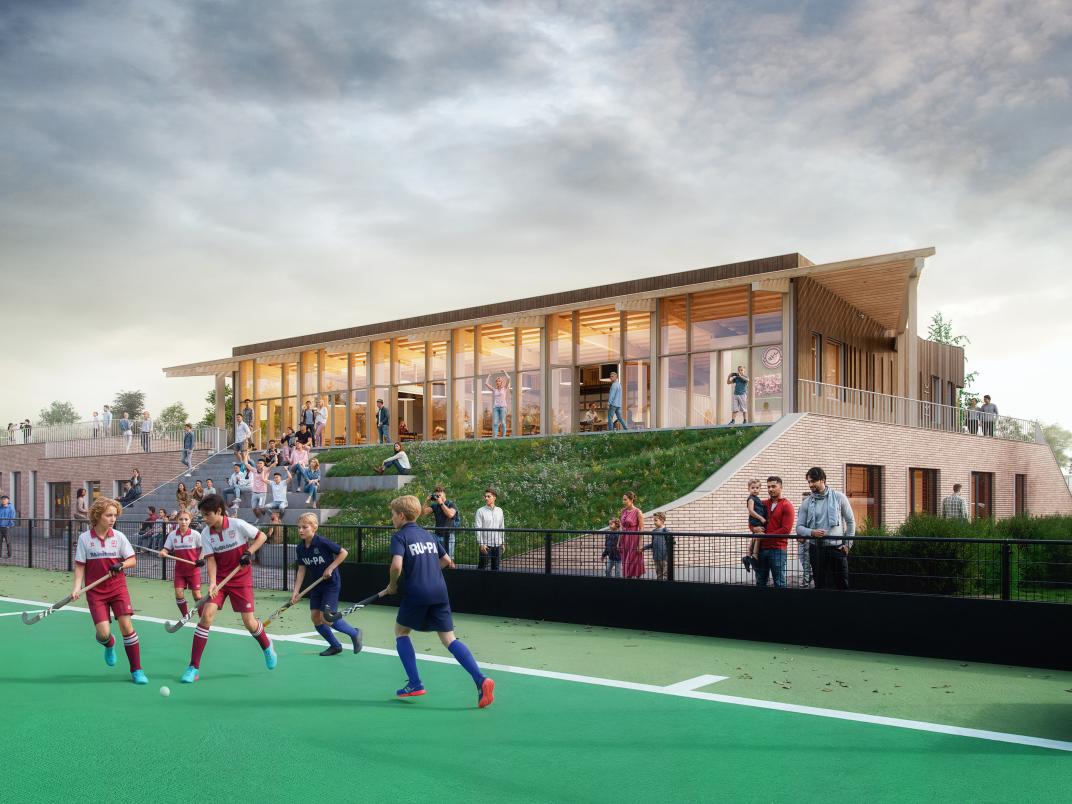 Sportveld bij MFA sportpark Elsenburg. Op het veld staan jongeren die hockeyen. Achter hen is een clubhuis te zien en naast het veld staan supporters.