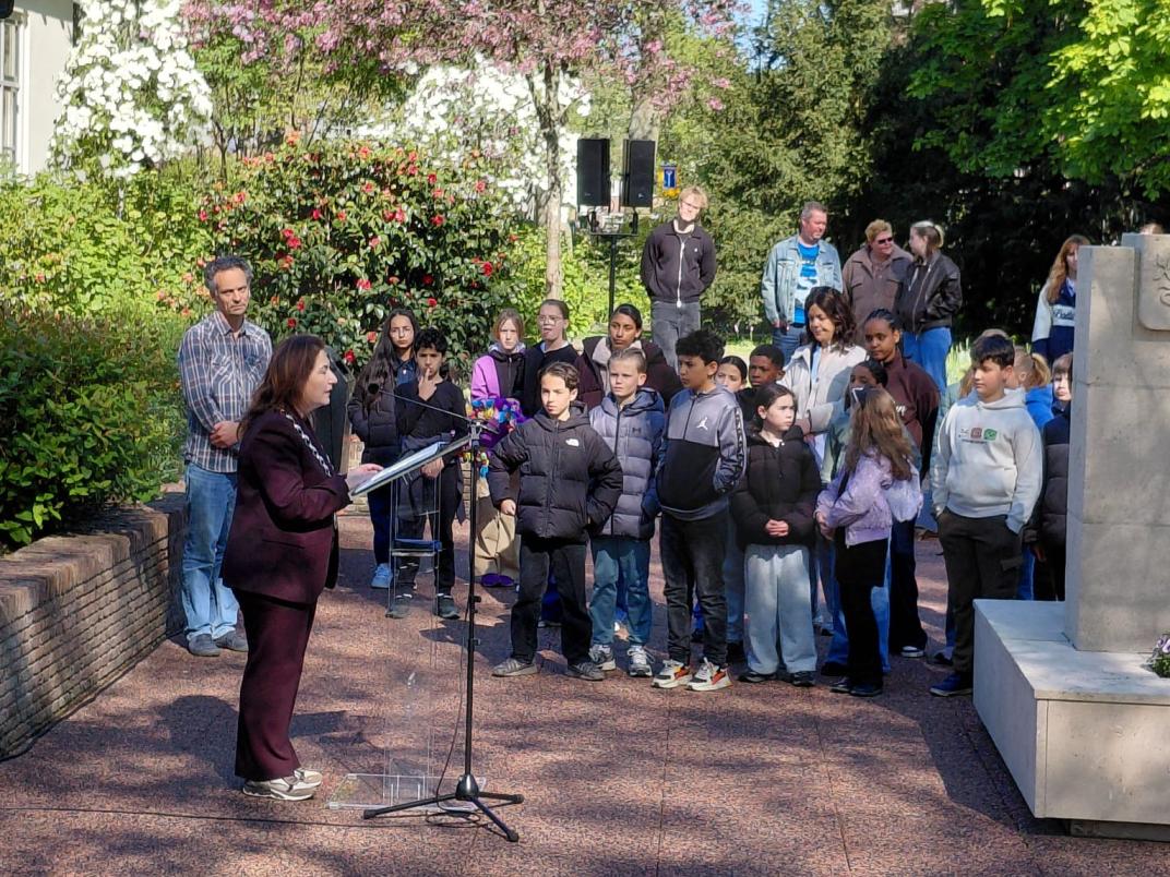 De burgemeester houdt een toespraak. Een groep kinderen staat aandachtig te luisteren naast het oorlogsmonument. 