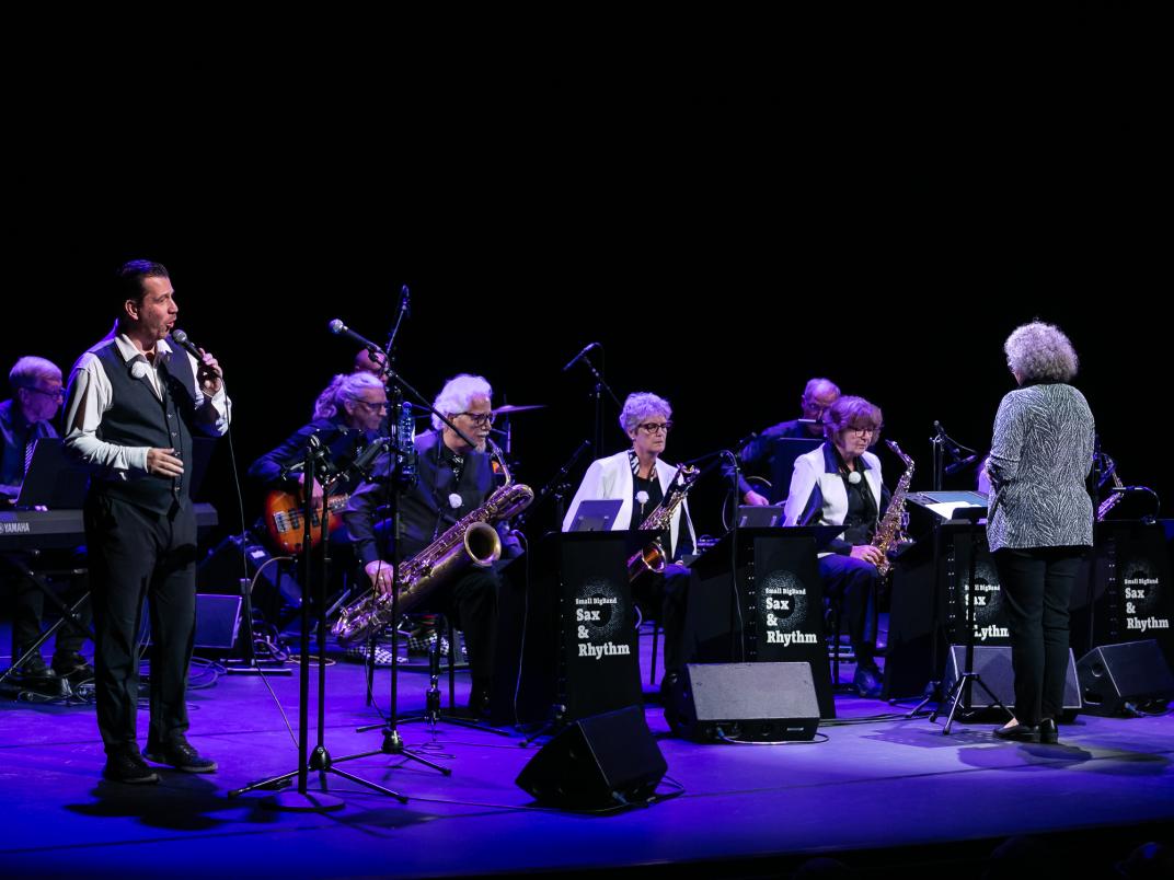 Overzichtsfoto van een orkest dat saxofoon bespeeld, een dirigent en zanger..